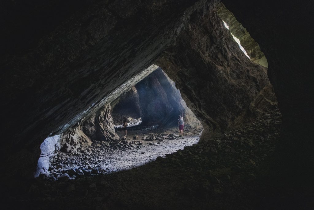 A picture of the insides of a big cave with a lot of light coming from the sides, filtering through big pillars of stone, with two people standing on the ground under the light