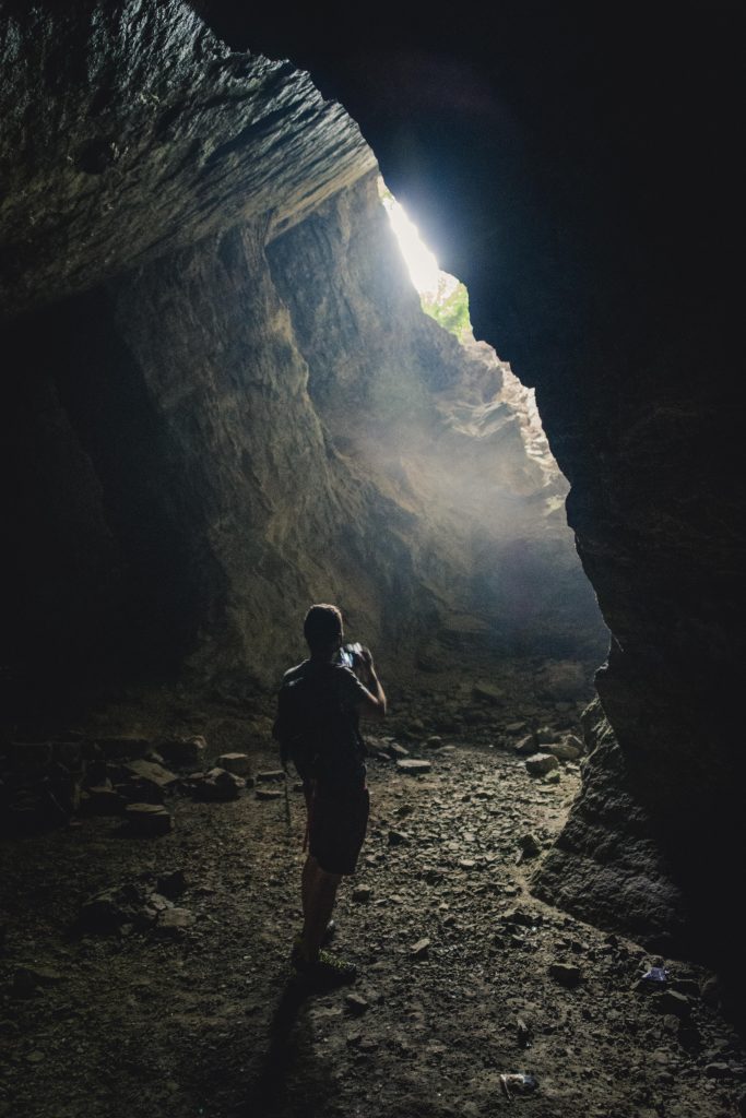 A guy taking a picture of the light coming into a dark cave, while standing inside the cave and looking at its exit