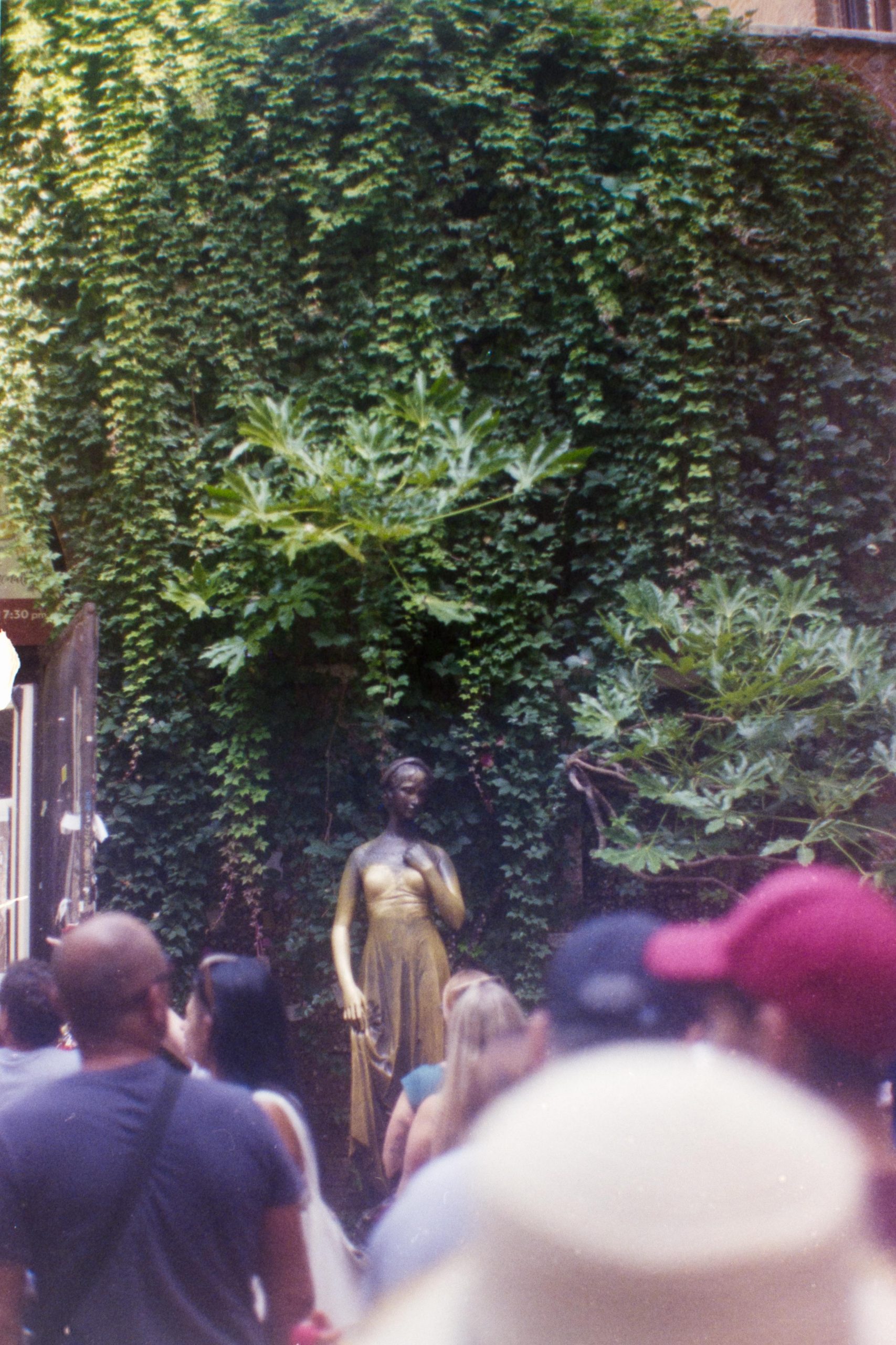 The golden statue of Juliet rising from a crowd in front of it with a wall full of plants behind