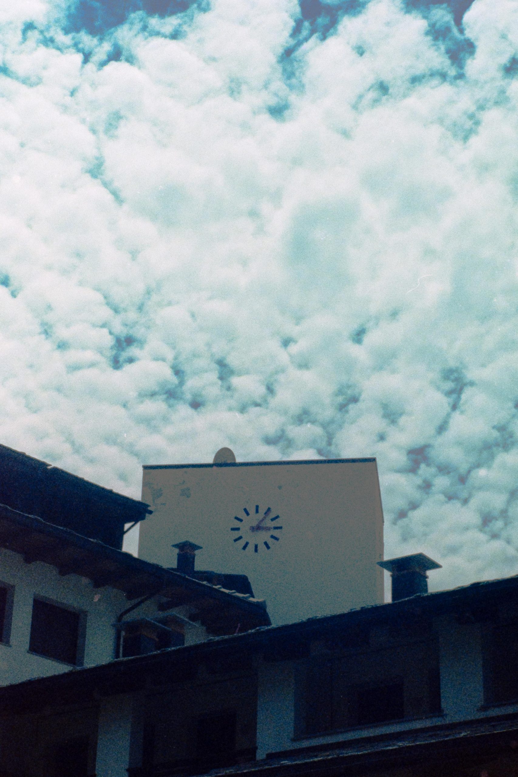 A minimalist clock tower behind some roofs with scattered clouds in the background