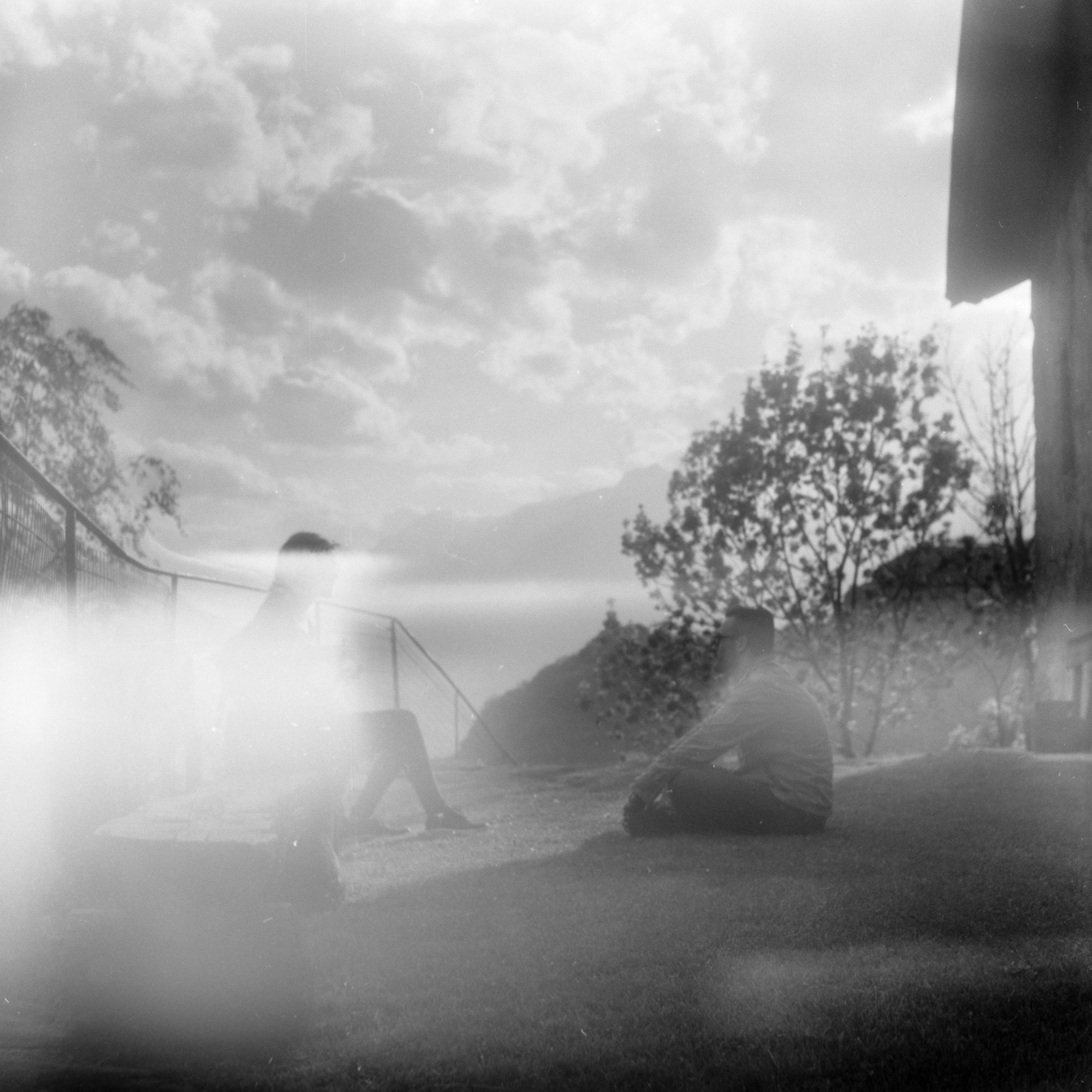 A guy sitting on the grass talking with a figure hidden by a light with clouds in the background