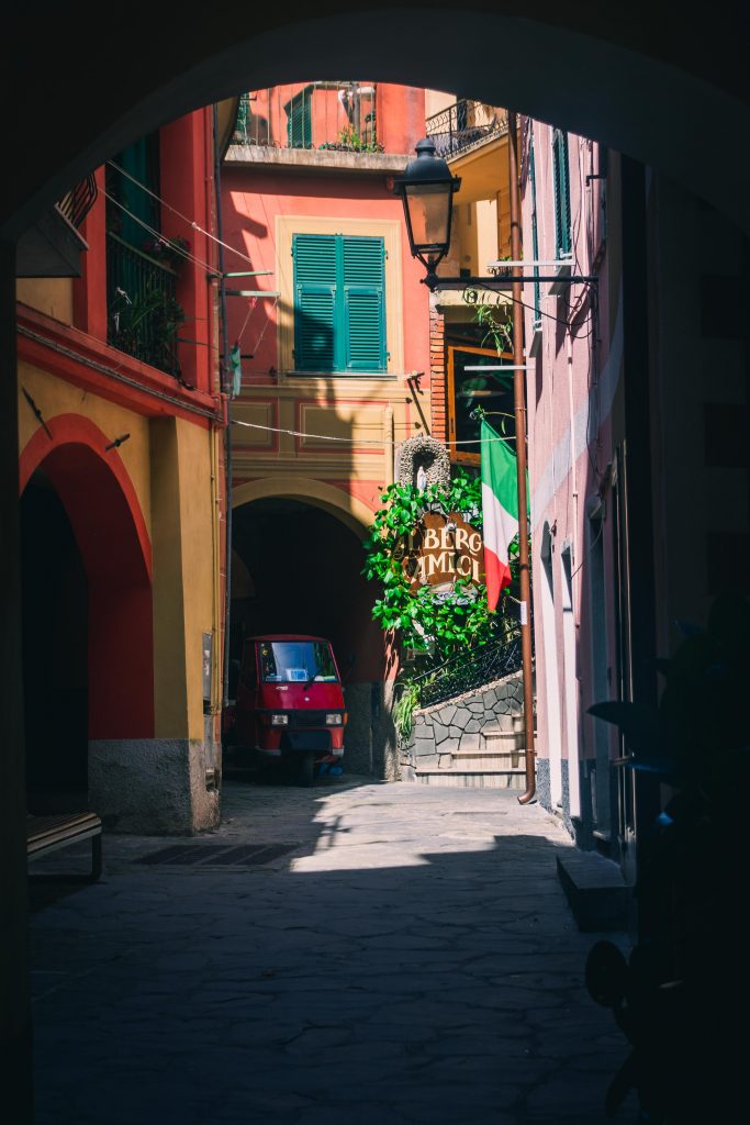 a red Ape parked in a colorful street with a sign citing "Albergo degli amici" on it